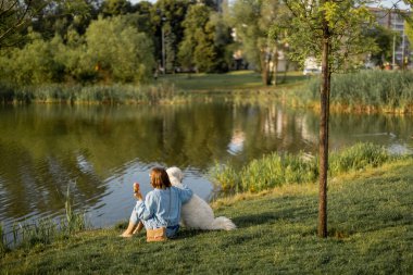 Genç bir kadın gölün kenarında sevimli beyaz köpeğiyle otururken dondurma yer. Geniş görüş. Evcil hayvanlarla arkadaşlık ve birlikte zaman geçirmek.