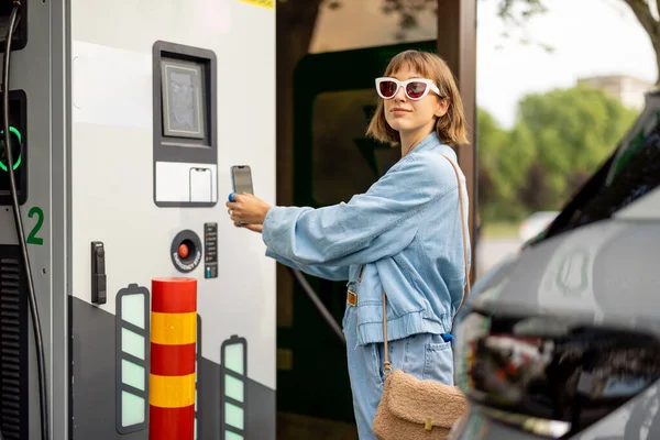 年轻女子打算在车用公共电站收取电动车费用 用电话扫描Qr代码支付费用 电动汽车充电现代技术的概念 — 图库照片
