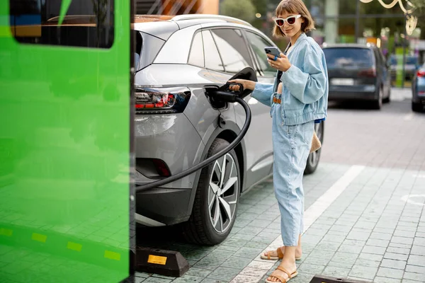 若い女性は屋外の公共充電ステーションで車の高速充電のためのケーブルにプラグ 電気自動車の充電 電気及び運転のための緑のエネルギーの概念 — ストック写真