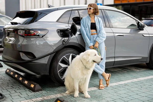 Nuori Nainen Jolla Valtava Valkoinen Koira Odottamassa Sähköauton Lataamista Julkisella — kuvapankkivalokuva