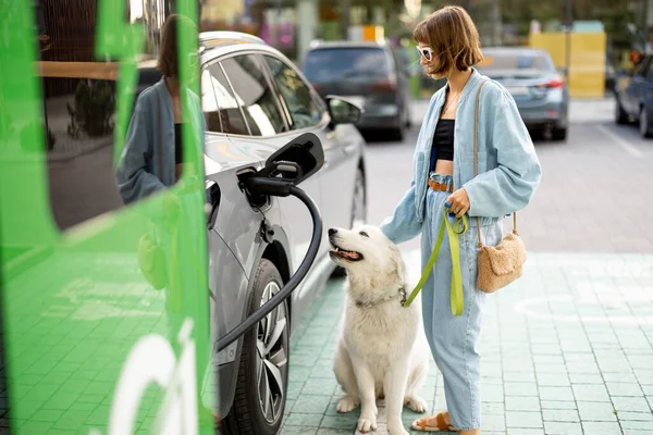 Happy woman with a huge white dog waiting for electric car to be charged on a public station outdoors. Concept of EV cars and friendship with pets