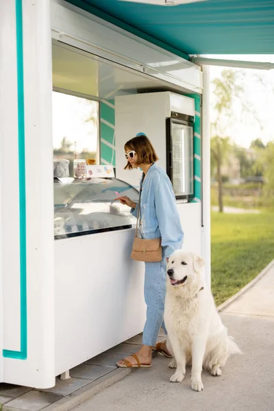 Young woman choosing ice cream to buy at beautiful shop while walking with her dog in a park. Spending summer time together with pet outdoors