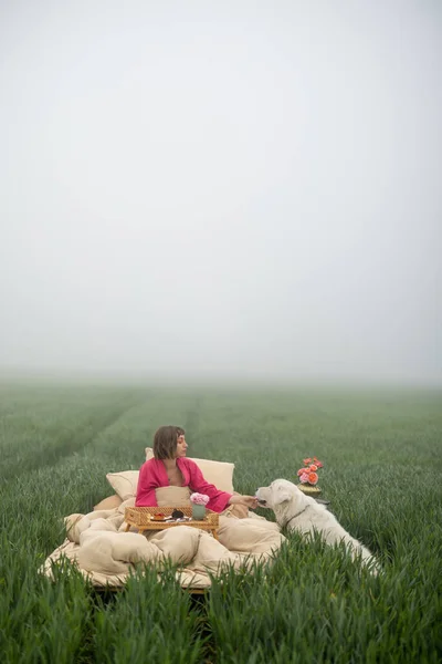 Young woman enjoys morning time, lying on bed with her cute dog and eating sweet breakfast on green field outdoors
