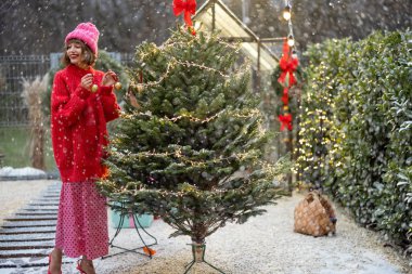 Kırmızılı genç kadın, kış tatiline hazırlanmak için evinin arka bahçesinde süslü balolar ve çelenklerle süslüyor.