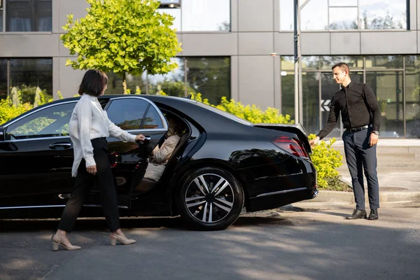 Female chauffeur opens a car door for a business lady going to sit, man packs a suitcase into a car trunk. Concept of luxury car transfer service, business trips and taxi