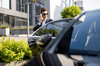 Ofis binasının yanında duran lüks arabanın yanında duran yakışıklı işadamı telefonla konuşuyor. İş hayatı ve ulaşım kavramı