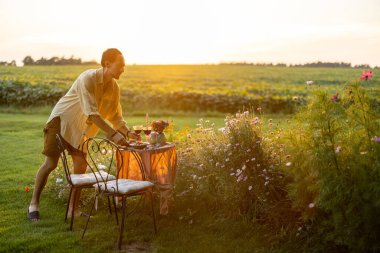 Gün batımında yeşil çimenlikte elinde çiçeklerle romantik bir akşam yemeği servisi yapan bir adam.