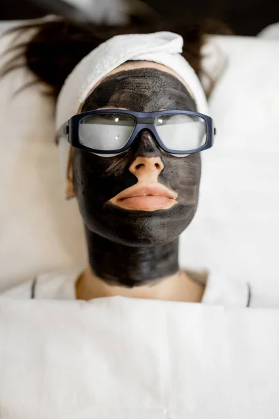 Young womans face in carbon mask and eyeglasses ready for laser carbon peeling at beauty salon, close-up. Concept of beauty and facial care