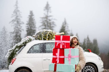 Kadın, Noel çelengi ve çatıda ağaçla süslenmiş arabanın yanında dikilmiş, karlı havada dağlarda arabayla seyahat ederken bir yığın hediye kutusu tutuyor. Kış tatili kavramı