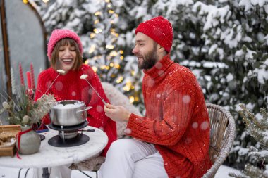 Man and woman have romantic dinner with fondue, while sitting together by the table at beautifully decorated snowy backyard. Young family celebrating winter holidays outdoors clipart