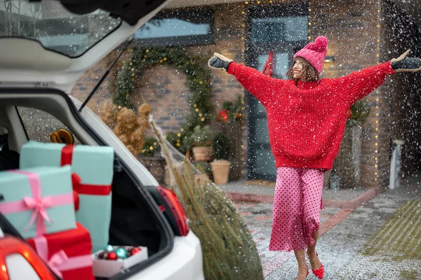 穿着红色毛衣的快乐女人来到一辆车上 车上有礼品盒和圣诞树 房子门廊上装饰着冬雪假期的装饰 为新年假期作准备的概念 — 图库照片