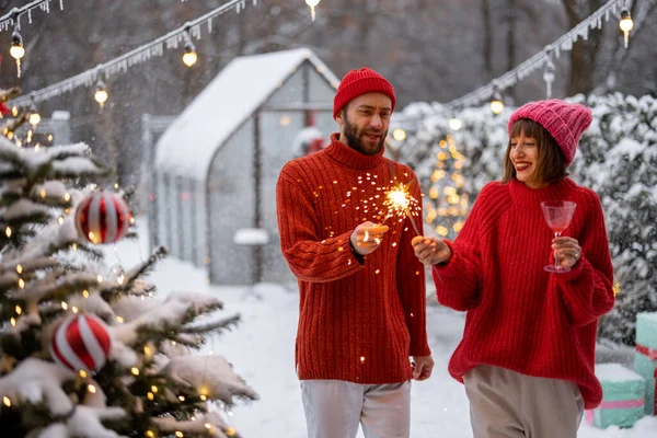 身穿红毛衣的男女们在雪地的后院点着火花 在圣诞树旁玩乐 庆祝新年 — 图库照片