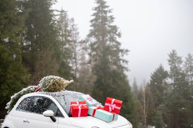Çatıda Noel ağacı olan beyaz bir araba. Karlı havada dağ yolunda hediyelerle süslenmiş. Yeni yıl anlayışı ve tatil hazırlığı.