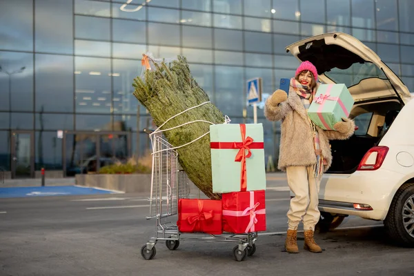一位快乐的年轻女子站在商场停车场旁的购物车旁 车上塞满了礼物和圣诞树 一边打电话一边说 — 图库照片