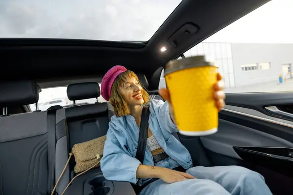 Stylish woman travels with coffee on backseat of a car, showing yellow coffee cup with blank space. Wide angle view, modern car with panoramic rooftop