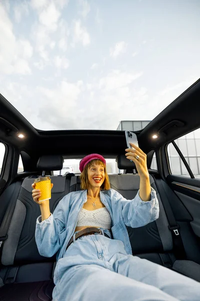 Young stylish woman enjoys traveling by car, sitting relaxed with a coffee cup and phone on backseat. Wide angle view, modern car with panoramic rooftop