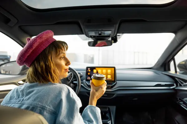 Stylish woman has a drink on the go while driving modern car at city. Concept of a coffee to go and travel by car