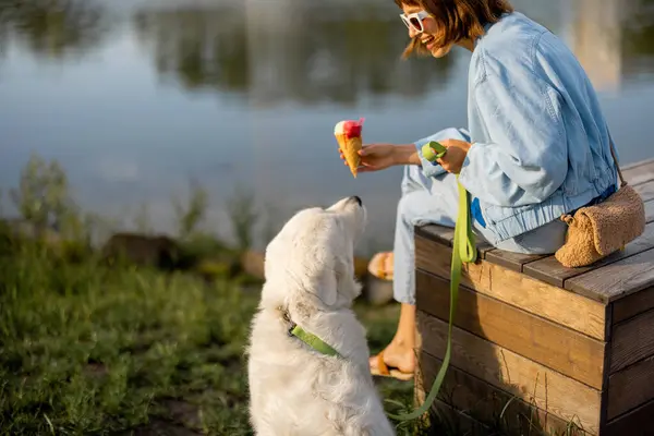 公園の湖の近くで可愛い白い犬と一緒に座っている間 若い女性はアイスクリームを食べます ペットとの友情と一緒に余暇を過ごす — ストック写真