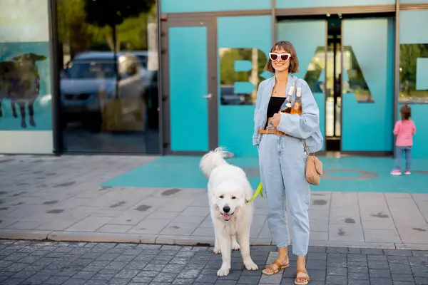Woman going with her cute white dog out of a supermarket, carrying wine bottles in front of a shop doors. Concept of firendship with pets and lifestyle