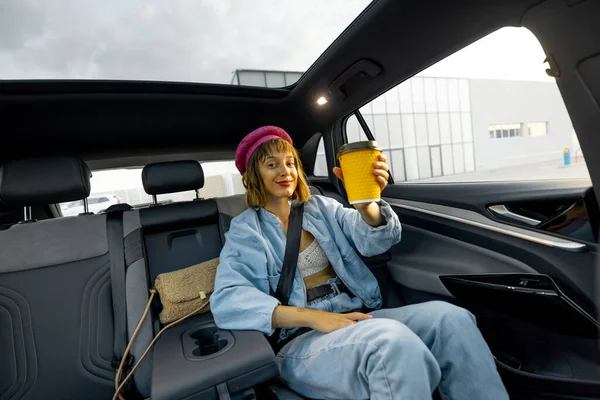 Stylish woman travels with coffee on backseat of a car, showing yellow coffee cup with blank space. Wide angle view, modern car with panoramic rooftop