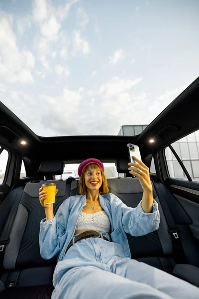 Young stylish woman enjoys traveling by car, sitting relaxed with a coffee cup and phone on backseat. Wide angle view, modern car with panoramic rooftop