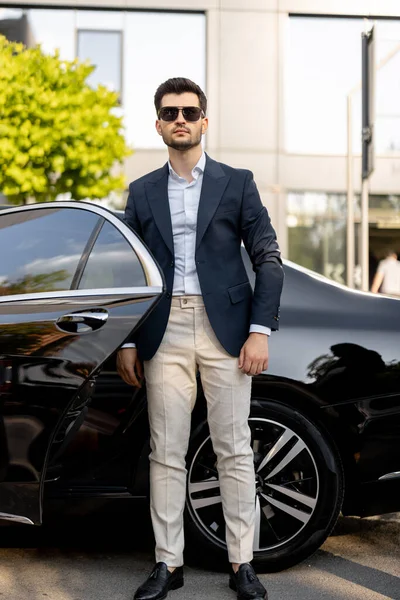 Portrait of a handsome businessman standing near luxury car outdoors. An elegant man gets out of a premium car