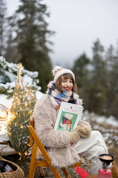 Young woman relaxes and enjoys calm on nature during winter holidays, sitting at picnic decorated for Christmas with a magazine in mountains