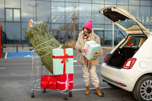 Young woman packing christmas presents and tree into car, standing with trolley at parking lot of a mall. Cheerful girl having festive shopping in anticipation of the winter holidays