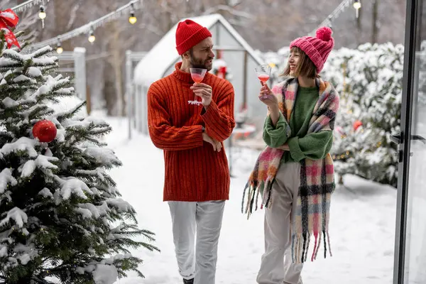 Man and woman talk and drink while spending winter time together at beautifully decorated snowy backyard. Young family celebrating Near Years holidays
