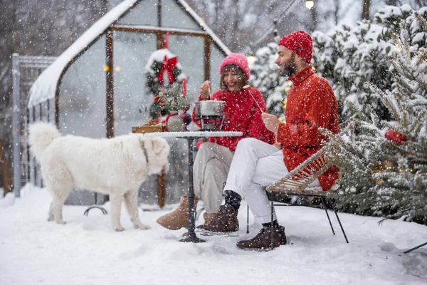 Man and woman have romantic dinner eating fondue and playing with dog, while sitting together by the table at beautifully decorated snowy backyard on winter holidays