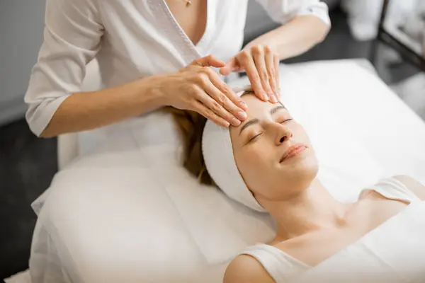 Beautician makes professional face massage for the womans face at beauty salon. Young adult caucasian woman receiving relaxing facial massage
