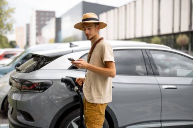 Dışarıdaki bir şarj istasyonunda telefonla beklerken şapkalı bir adam elektrikli aracın kablolarını fişe takıyor. Elektrikli araba ve yeşil enerji ile seyahat kavramı