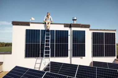 Üzerinde güneş enerjisi santrali olan özel bir evin çatısı, tepesinde oturan ve mülkünün tadını çıkaran bir adam. Yenilenebilir enerji ve sürdürülebilir yaşam tarzı kavramı