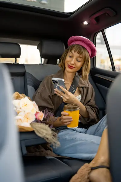 Nuori Tyylikäs Nainen Käyttää Älypuhelinta Istuessaan Kahvikupin Kanssa Auton Takapenkillä — kuvapankkivalokuva