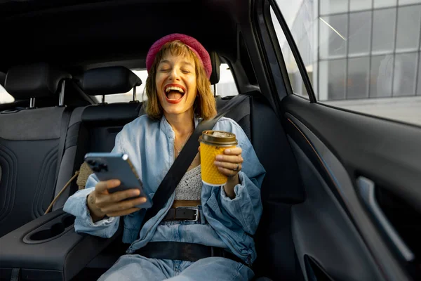 Iloinen Nainen Käyttää Älypuhelinta Istuessaan Rento Kahvikuppi Takapenkillä Auton Liikkeellä — kuvapankkivalokuva