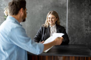 Genç bir tıbbi resepsiyonist erkek bir müşteriyle konuşuyor ve modern kliniğin resepsiyonunda imzalaması için bir kaç kağıt veriyor.