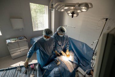 Ameliyat tedavisi devam ediyor. İki cerrah, bir hastanın alt karın bölgesini ameliyat ediyor. Yukarıdan görünüyor.
