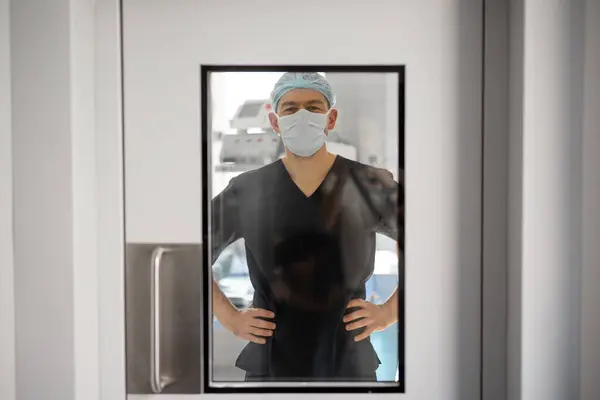 Portrait of a surgeon looks through a glass door while standing in operation room. Concept of surgery