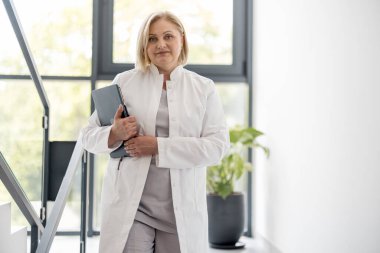 Klinikteki merdivenlerde, medikal önlüklü orta yaşlı hemşire portresi. Kapalı alanda klasörü olan güzel beyaz bir sağlık görevlisi.