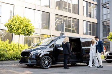 Female chauffeur waits a business people to let them in a minivan taxi, keeping door open. Concept of business trips and transportation service clipart