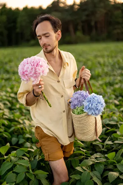 Handsome man walks with flowers on farmland outdoors. Young farmer or florist on green field during sunset