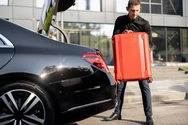 Chauffeur of a business-class car packs red suitcase into a trunk on the street near an office building. Concept of business trips, personal driver or luxury taxi