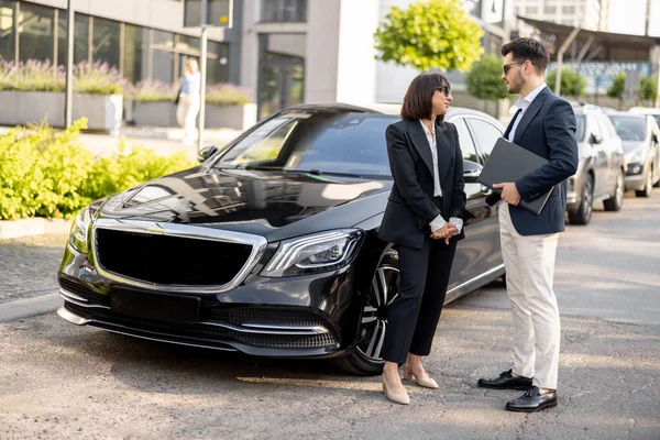 Business partners have a conversation while standing together near luxury car outdoors