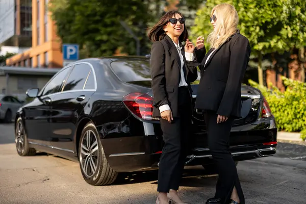 Full body portrait a two women in formal wear as a drivers of luxury taxi standing near car on a street. Concept of business transfer services, chauffeur portrait or business trips