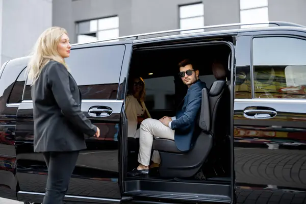 Female chauffeur helps a business man to get out of vehicle, opening a door of luxury black minivan. Concept of personal driver, luxury taxi for business delegation