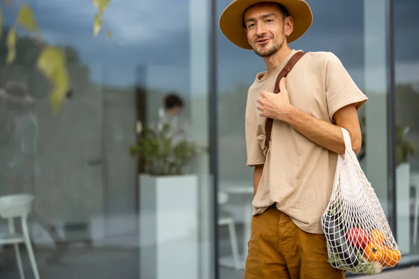 Portrait of a stylish man in hat walks with a mesh bag full of fresh vegetables near supermarket outdoors. Concept of sustainable lifestyle and style