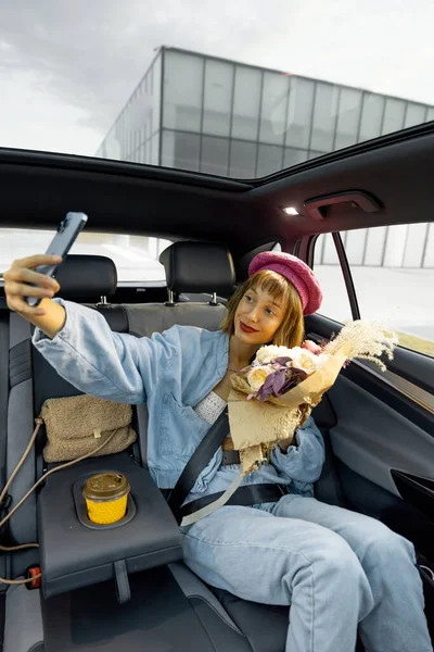 Young stylish woman makes selfie on phone or has a video call, while sitting happily with flowers on backseat of a car. Woman traveling by car at city. Wide view, vehicle with panoramic rooftop
