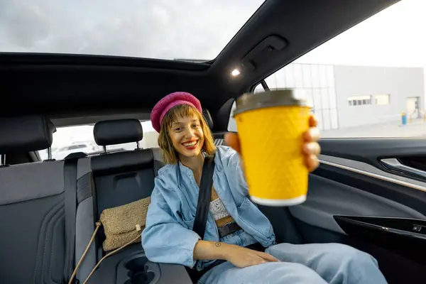 スタイリッシュな女性は 車の後部座席にコーヒーと一緒に旅行し 空白のスペースで黄色いコーヒーカップを示しています 広角ビュー パノラマ屋上モダンな車両 ロイヤリティフリーのストック写真