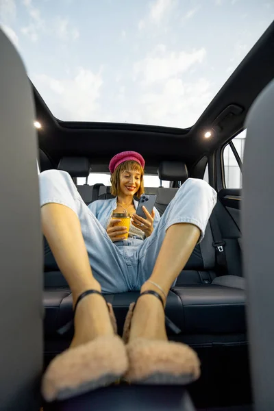 若いスタイリッシュな女性は バックシートでコーヒーカップと電話でリラックスして車で旅行を楽しんでいます 広角ビュー パノラマ屋上モダンな車両 ロイヤリティフリーのストック画像