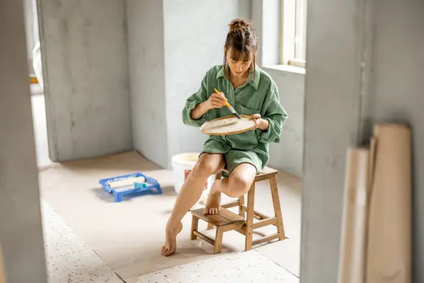 若い女性は新しいアパートの修理中に壁を塗ります 椅子に座り 塗料の色を選択する ホームリフォームと修理コンセプトのクリエイティブプロセス ロイヤリティフリーのストック写真